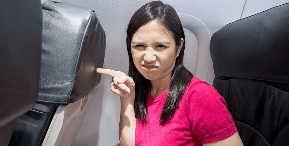 Женщина в самолете с недовольным выражением лица указывает на кресло впереди, показывая возмущение поведением соседки