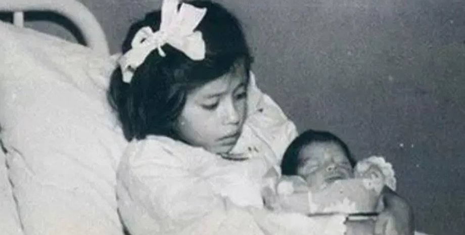 Девочка сидит на больничной кровати и держит на руках новорожденного ребенка, завернутого в одеяло