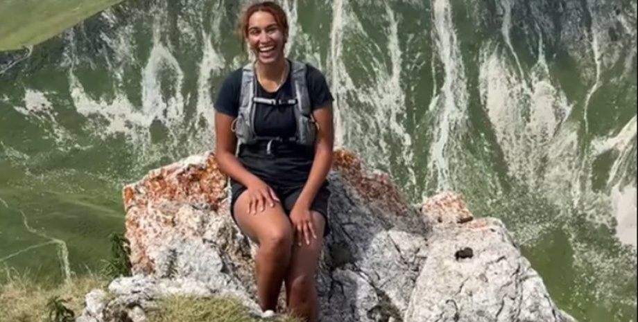 Молодая женщина улыбается, сидя на скале, рядом с ней ходят дружелюбные козы