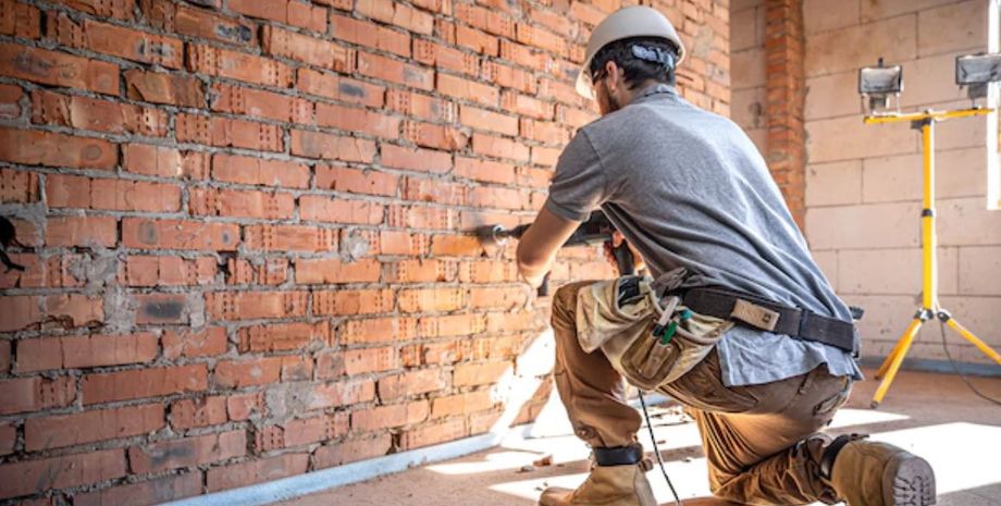 Мужчина в строительной каске и с инструментами вскрывает кирпичную стену во время ремонта