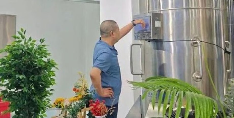 Мужчина в голубой рубашке стоит у большой металлической криокамеры и нажимает на панель управления