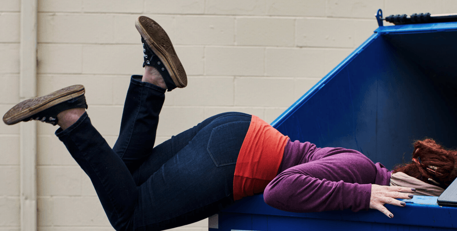 Женщина в яркой одежде наклоняется в мусорный контейнер, заглядывая внутрь в поисках выброшенных вещей