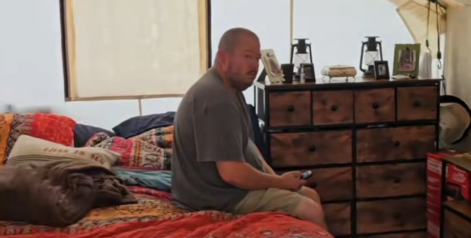 Мужчина сидит на кровати внутри брезентовой палатки с деревянным комодом и домашним интерьером