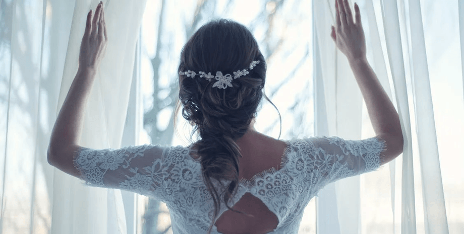 Женщина в кружевном свадебном платье стоит у окна, глядя во двор