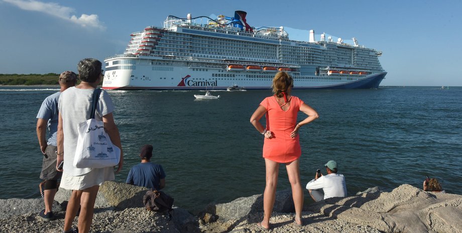 Люди стоят на берегу и смотрят на огромный круизный лайнер Carnival, уходящий в море